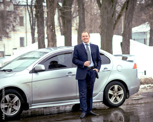A business man in a strict blue suit by a car on the road in winter. Transport and auto. Business. Copy space on the left © Alina