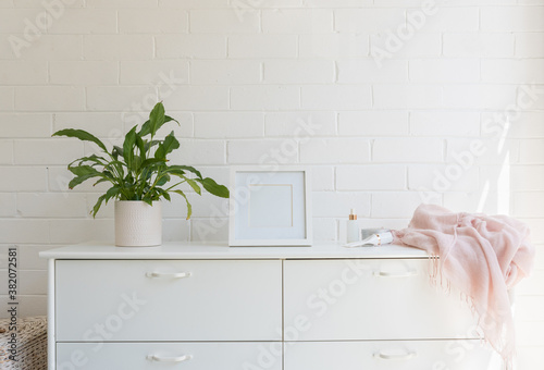 Close up of white dresser against painted brick wall with pot plant, blank square frame, pink scarf and lotions (selective focus) photo
