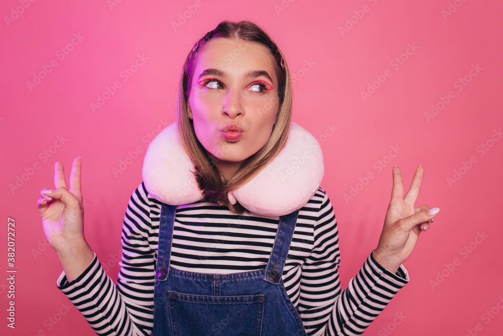 Playful pretty young woman in casual clothes stands over pink background, her lips in a tubule, makes peace or victory signs with both hands. White travel pillow on her neck. Playful pretty young woman in casual clothes stands over pink background, her lips in a tubule, makes peace or victory signs with both hands. White travel pillow on her neck.