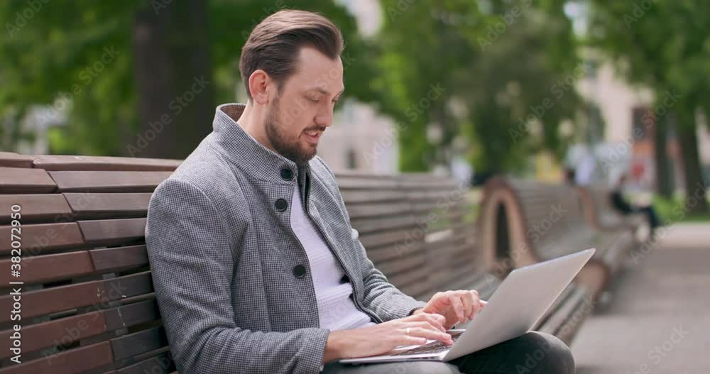 Young man with mustaches and a beard sitting on a bench in the square, leaning on the back of the bench, typing on the laptop keyboard. Green trees and grass, buildings and wooden benches are on the Young man with mustaches and a beard sitting on a bench in the square, leaning on the back of the bench, typing on the laptop keyboard. Green trees and grass, buildings and wooden benches are on the