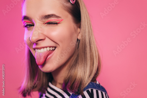 Very emotional pretty young woman in white and black striped sweatshirt winking and showing a tongue. Attractive happy caucasian woman. Pink stido background. Playful mood concept.