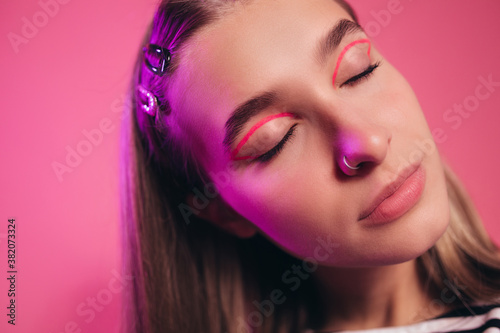 Nice charming relaxing girl with dreamy expression on her face closed her eyes. Close-up cut portrait of girl with bright pink cat-eye makeup  light brown hair with cute hair clips. Pink background.