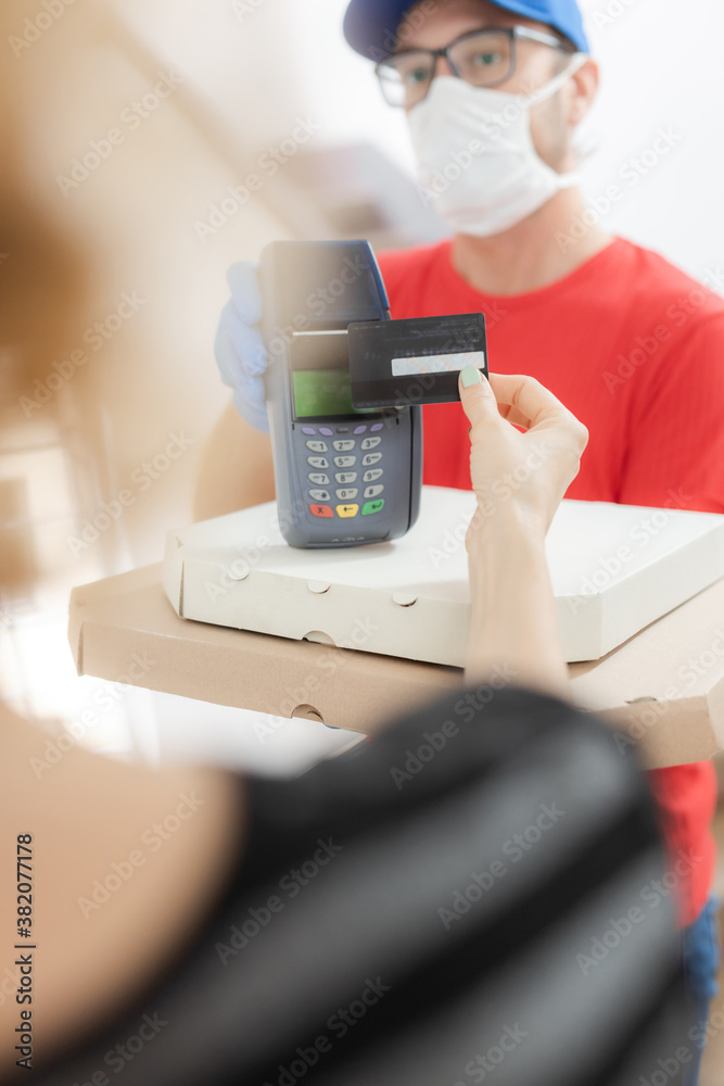 Delivery guy with protective mask holding pizza box and contactelss credit card terminal at the door. Delivery guy with protective mask holding pizza box and contactelss credit card terminal at the door.