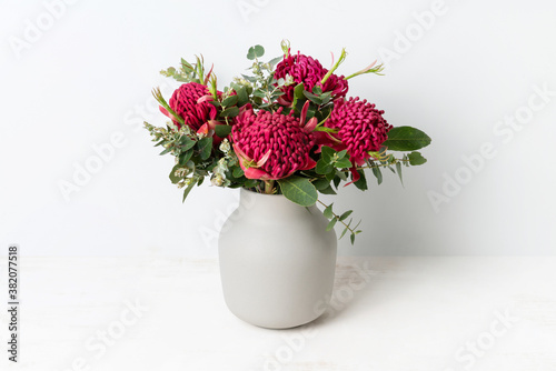 Fototapeta Naklejka Na Ścianę i Meble -  A bunch of Australian native red waratah flowers and eucalyptus leaves, in a stylish ceramic grey vase on a table, with a white background.