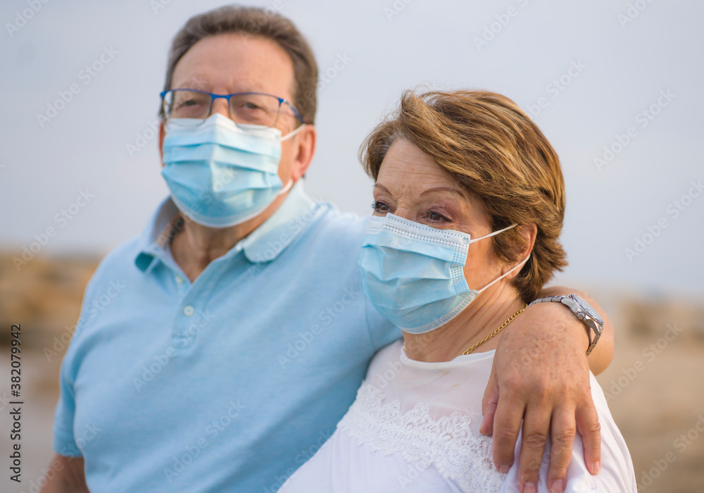 pensioner woman and her husband taking romantic walk together - happy retired mature couple in face mask walking on the beach in new normal holidays trip during covid19