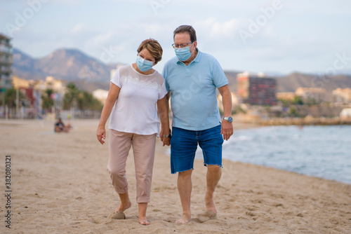 pensioner woman and her husband taking romantic walk together - happy retired mature couple in face mask walking on the beach in new normal holidays trip during covid19