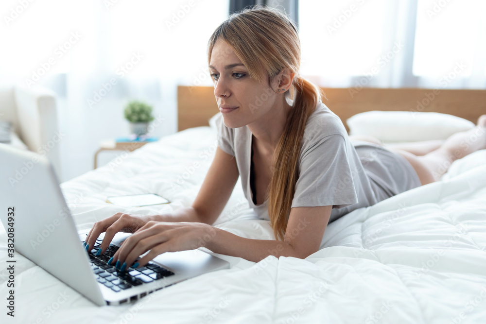 Shot of young beautiful woman using her computer while lying on the bed at home. Shot of young beautiful woman using her computer while lying on the bed at home.