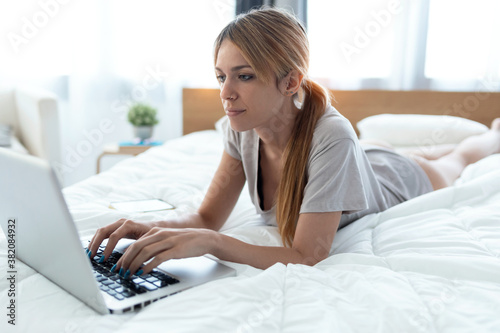 Shot of young beautiful woman using her computer while lying on the bed at home.