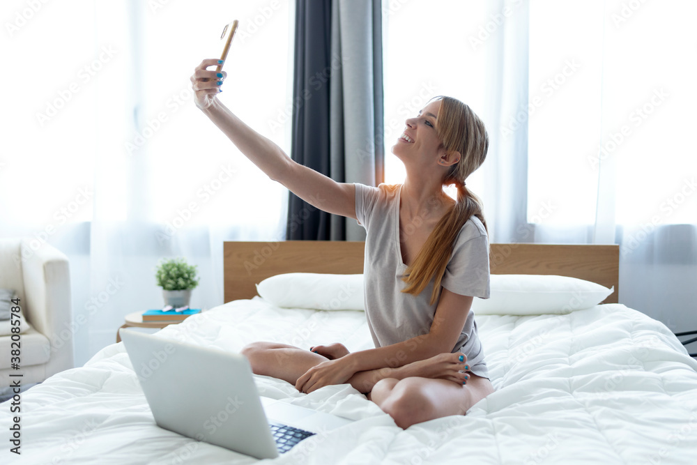 Happy young woman making selfie with her smart phone while working with laptop sitting on the bed at home. Happy young woman making selfie with her smart phone while working with laptop sitting on the bed at home.