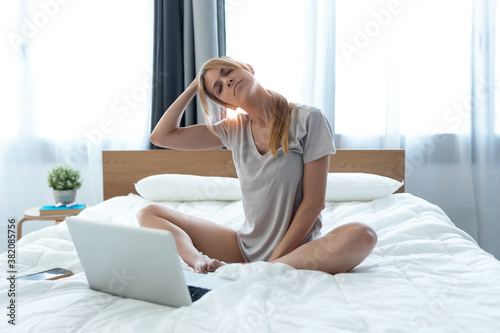 Beautiful young woman with neck pain working with laptop sitting on the bed at home.