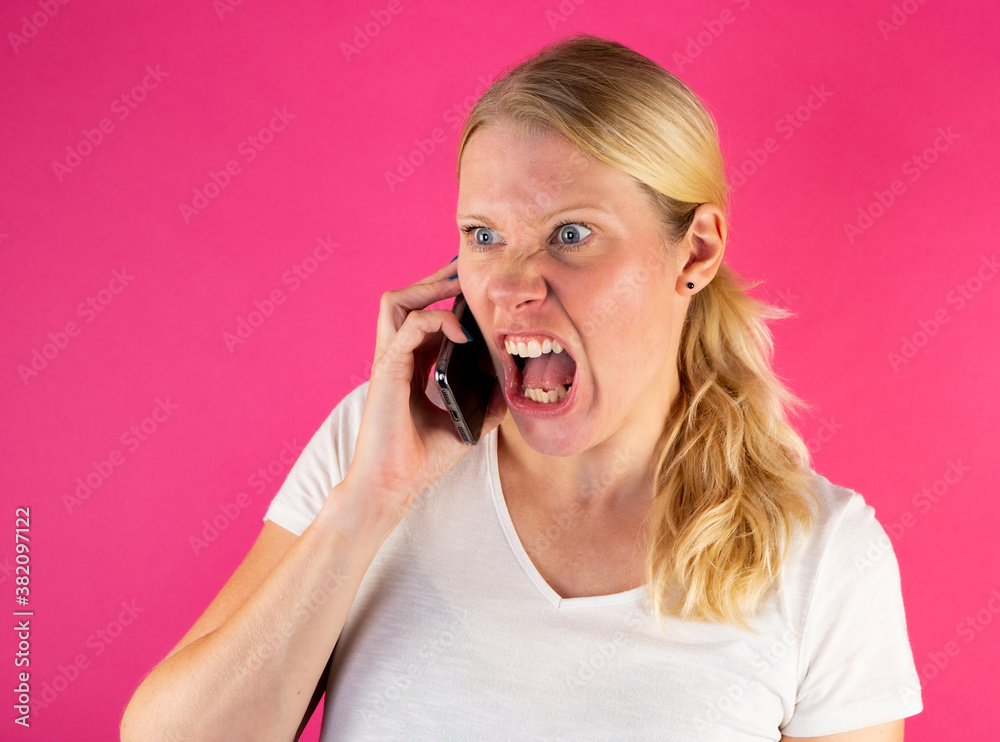 Blonde young woman screaming while making a phone call on smartphone. Isolated on pink background. Blonde young woman screaming while making a phone call on smartphone. Isolated on pink background.