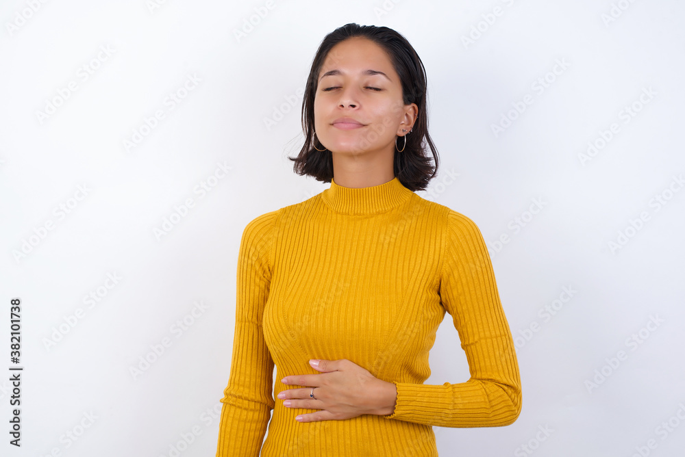 Young hispanic girl with short hair wearing casual yellow sweater isolated over white background touches tummy, smiles gently, eating and satisfaction concept. Young hispanic girl with short hair wearing casual yellow sweater isolated over white background touches tummy, smiles gently, eating and satisfaction concept.
