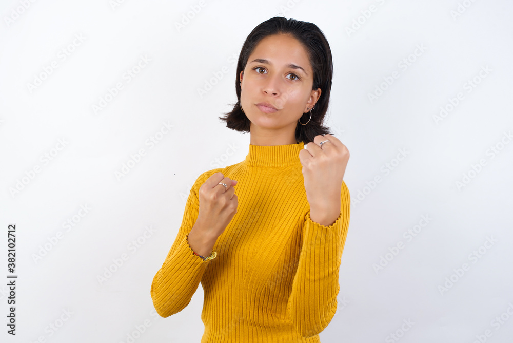 Young hispanic girl with short hair wearing casual yellow sweater isolated over white background Ready to fight with fist defense gesture, angry and upset face, afraid of problem. Young hispanic girl with short hair wearing casual yellow sweater isolated over white background Ready to fight with fist defense gesture, angry and upset face, afraid of problem.