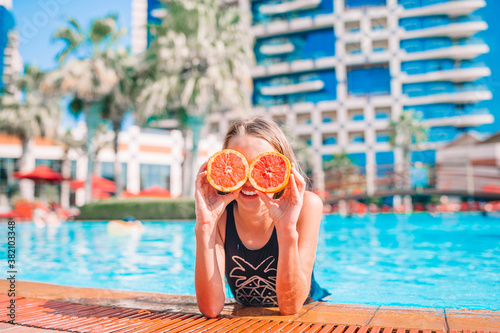 Little girl covering eyes with orange halves near eyes on background swimming pool