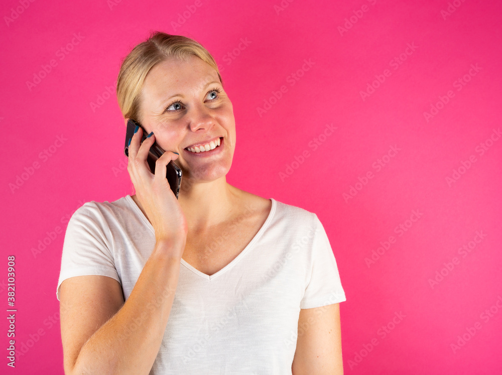 Blonde young woman smiling and looking up while making a phone call on smartphone. Isolated on pink background. Blonde young woman smiling and looking up while making a phone call on smartphone. Isolated on pink background.