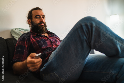 Bearded man sits on a couch while using the tv remote controller