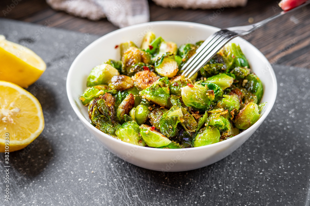 Woman or man eating roasted / fried brussels sprouts from a white bowl with chili and garlic flakes seasoning using a fork. Lemon halves and towel sitting on a cutting board in the background.