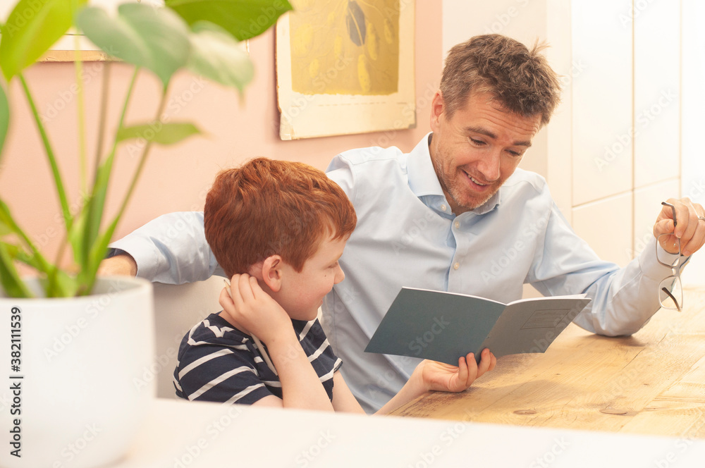 Candid picture of a smiling dad helping pre-teen boy with his school work at home during homeschooling, due to the coronavirus lockdown. Landscape format. Candid picture of a smiling dad helping pre-teen boy with his school work at home during homeschooling, due to the coronavirus lockdown. Landscape format.