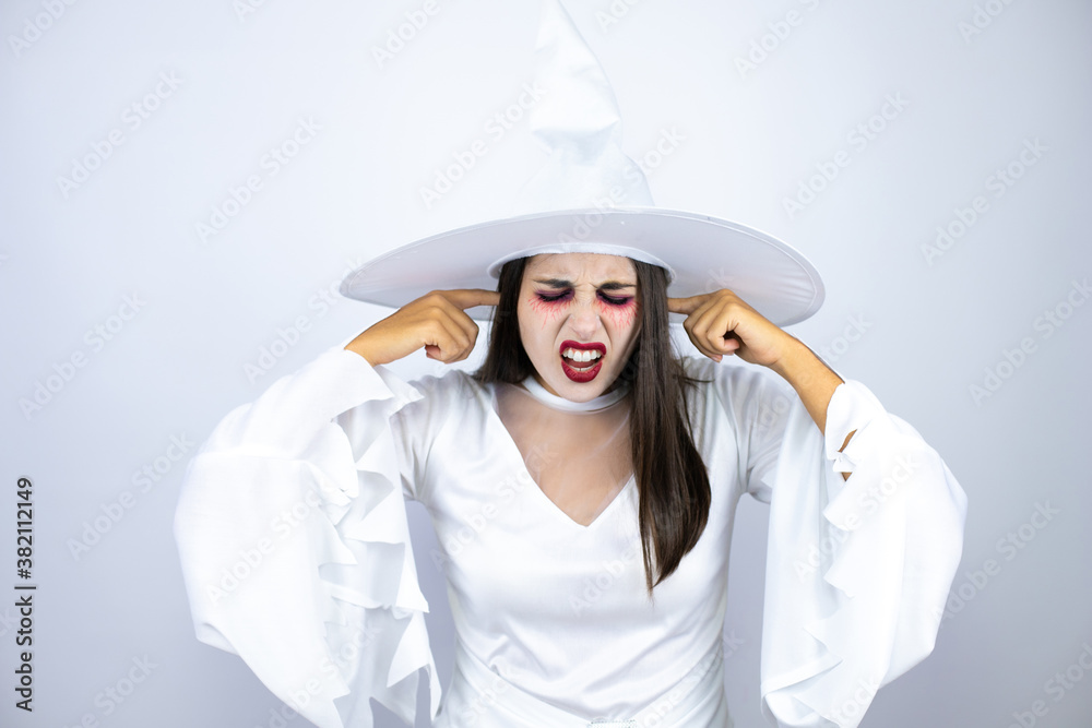 Woman wearing witch costume over isolated white background covering ears with fingers with annoyed expression for the noise of loud music. Deaf concept. Woman wearing witch costume over isolated white background covering ears with fingers with annoyed expression for the noise of loud music. Deaf concept.