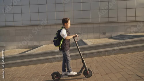 School boy riding with his electric scooter in the city with backpack on sunny day. Child in colorful clothes biking on way to school. Electric transport.