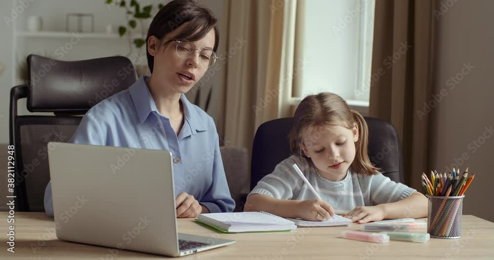 Portrait of two female persons, young smiling woman uses computer for presentation in lesson with little cute preschool girl, shows her video or photo, teaches to write and read, sit together at table Portrait of two female persons, young smiling woman uses computer for presentation in lesson with little cute preschool girl, shows her video or photo, teaches to write and read, sit together at table