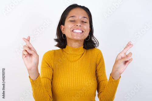Young hispanic girl with short hair wearing casual yellow sweater isolated over white background gesturing finger crossed smiling with hope and eyes closed. Luck and superstitious concept.