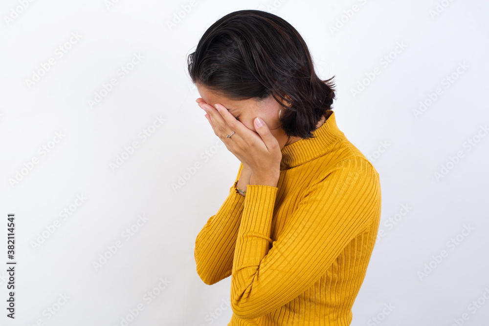 Sad Young hispanic girl with short hair wearing casual yellow sweater isolated over white background crying covering her face with her hands. Sad Young hispanic girl with short hair wearing casual yellow sweater isolated over white background crying covering her face with her hands.