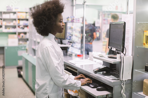 Smiling African American woman clinical pharmacist typing on computer keyboard, while standing behind the counter of modern hospital pharmacy