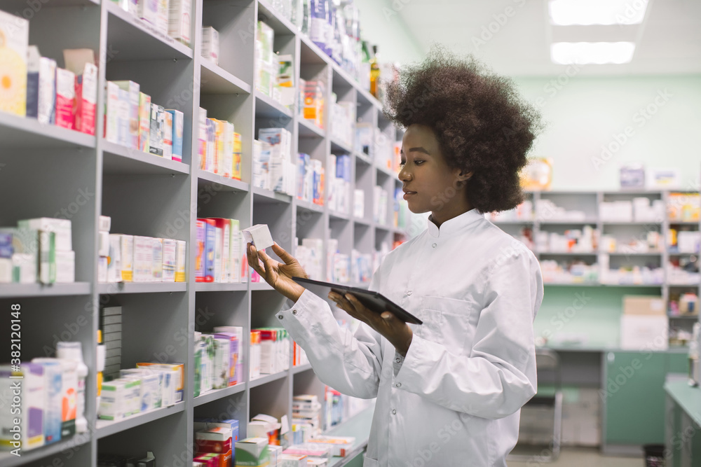 Young African American woman pharmacist or chemist using digital tablet, while standing near the shelves with medicines in modern pharmacy during drugs inventory