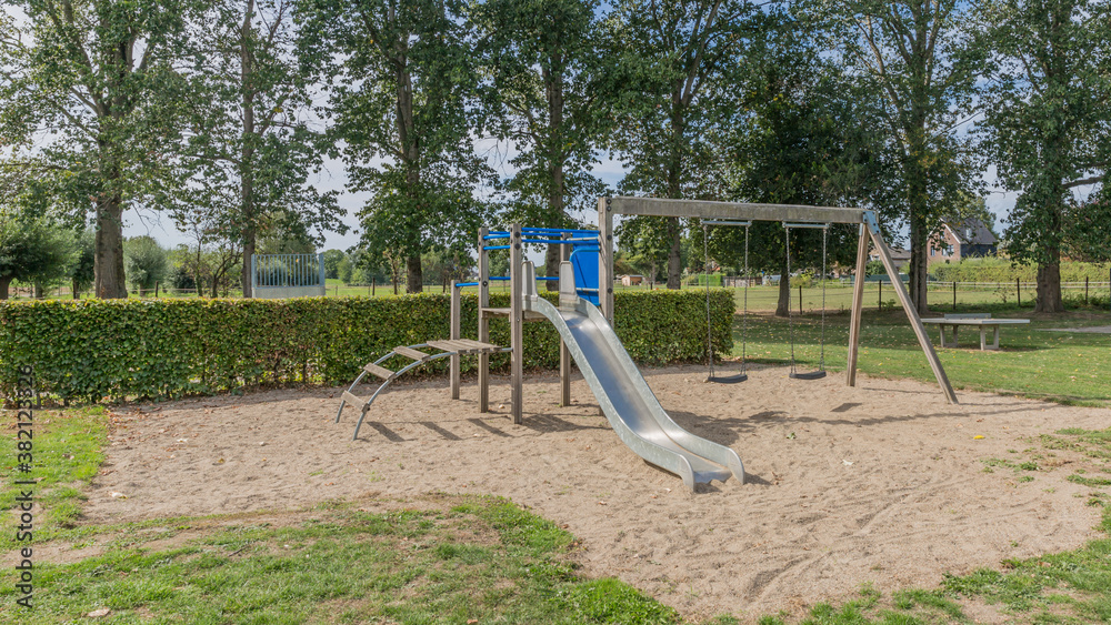 Outdoor games with swings and a slide in the playground on a sandy area of ​​a park surrounded by bushes and trees, sunny day to enjoy outdoor activities in the Netherlands Holland