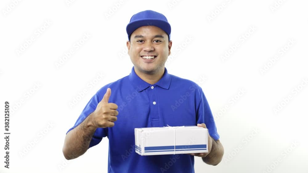 Smiling young asian delivery man in blue uniform showing thumbs up holding parcel cardboard box on isolated white background. 4k Resolution.