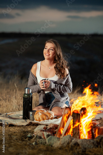 Beautiful girl drinking tea by the fire. Pcnic rest. Thermos tea. Romantic atmosphere in nature. Campfire. Bonfire