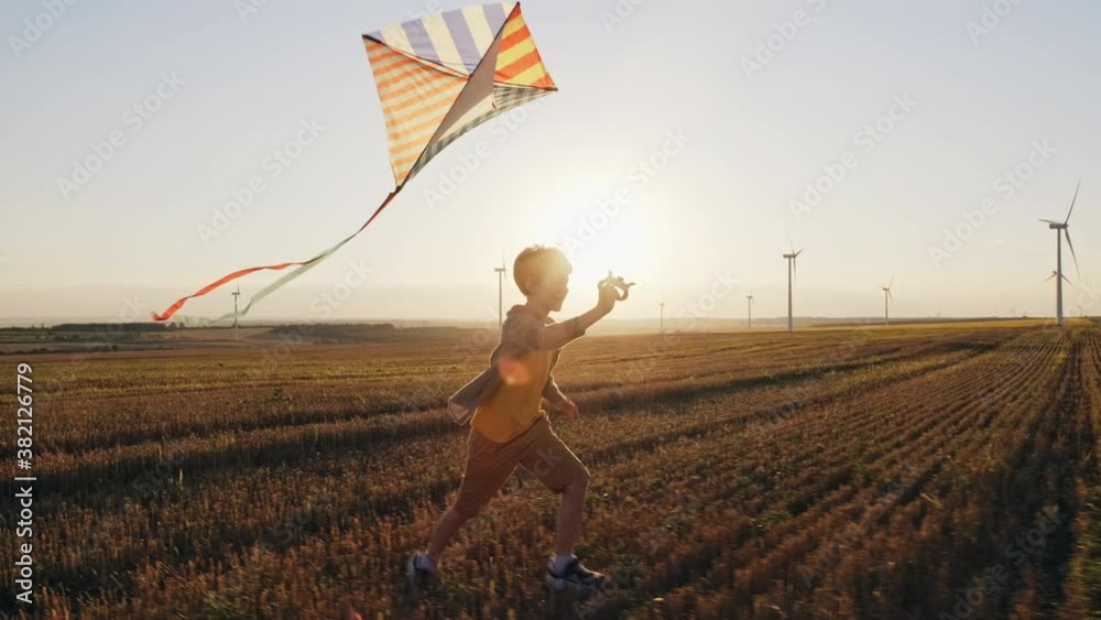 Happy boy runs launches bright kite into sky mown wheat field, playing with wind in field of an orange sunset on day lens flares wind turbines in summer slow motion. School break. Lifestyle. Childhood