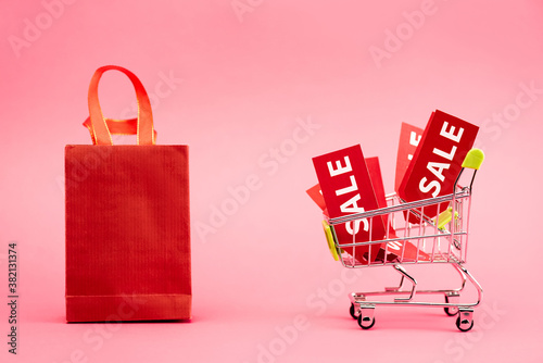 red tags with sale lettering in shopping trolley near paper bag on pink