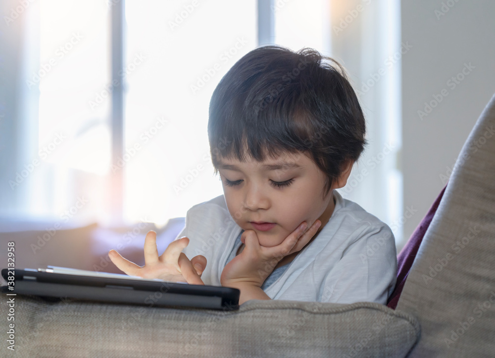 Kid touching finger on tablet screen, Child boy lying on sofa relaxing at home watching cartoons or playing games on digital pad, Home schooling, Social Distance, E-learning online education Kid touching finger on tablet screen, Child boy lying on sofa relaxing at home watching cartoons or playing games on digital pad, Home schooling, Social Distance, E-learning online education