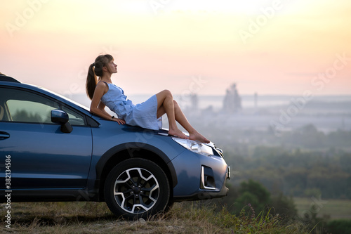 Happy woman driver in blue summer dress enjoying warm evening laying on her car hood. Travel and vacations concept.