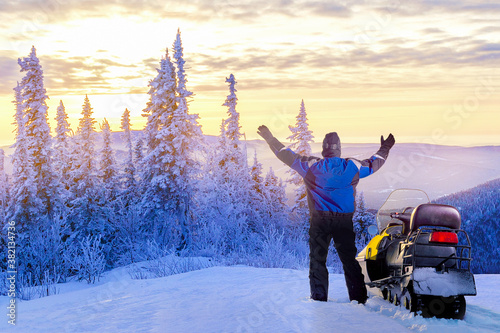  Man standing on snowy mountain near snowmobile enjoying view and achievement on sunset winter day.