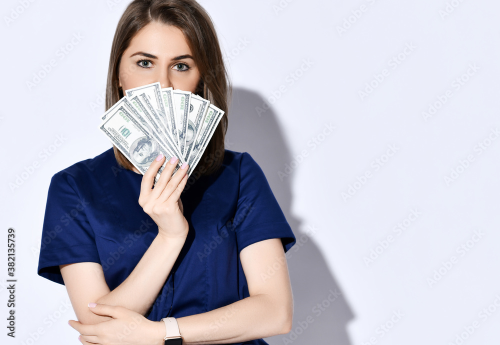 Portrait of young beautiful woman dentist, doctor, nurse holding fan of dollars cash bills in front of her face Portrait of young beautiful woman dentist, doctor, nurse holding fan of dollars cash bills in front of her face