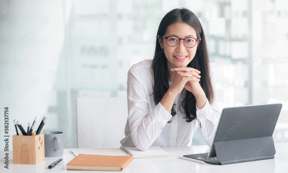 Portrait of young attractive asian woman with glasses relaxing while sitting at her office desk, smiling and looking at camera. - obrazy, fototapety, plakaty 