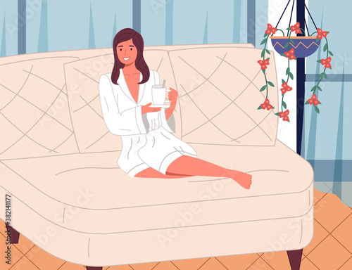 Young beautiful smiling woman sitting with bent legs on a large white comfortable sofa with cup of tea or drink, white bathrobe. Girl in the spa. Beauty treatments and relaxation. Cozy interior