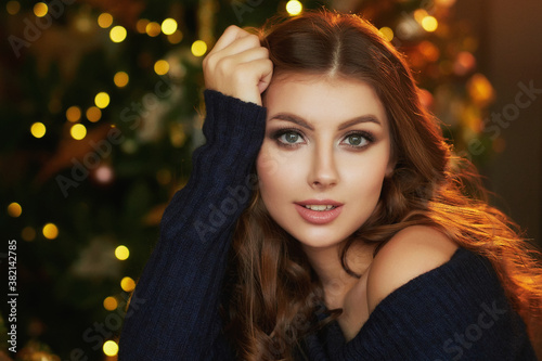 Close up portrait of a young beautiful smiling woman posing in an interior with festive Christmas lights on the background.