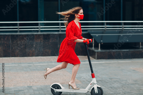 Young woman with electric scooter in red dress and gloves with face mask at the city. New normal fashion and coronavirus COVID protection concept.