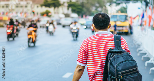 Back view of male patient with mask in red and white shirt with a black backpack standing at bus stop and waving his hand for Taxi or Bus in the city to go to the hospital.