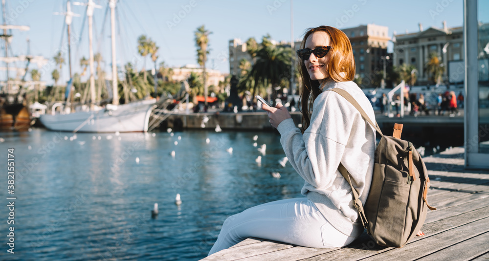 Portrait of cheerful hipster girl in stylish sunglasses resting near harbor during summer vacations holding cellphone for blogging and smiling at camera, happy woman with backpack enjoying leisure