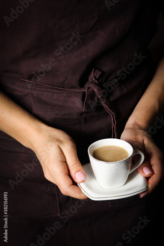 Coffee with cream in a white cup in the hands of a waiter in a black apron. Background image  copy space