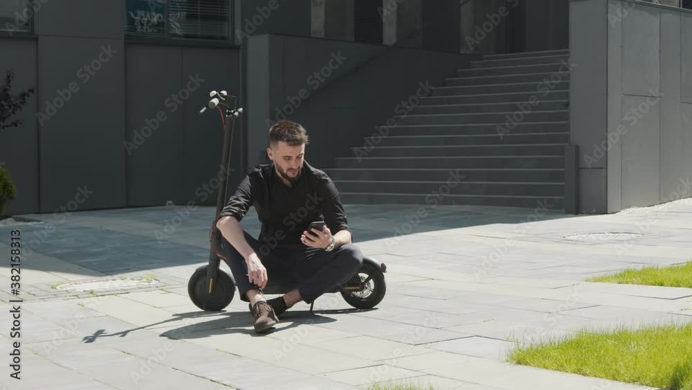 Male student sitting on an electric scooter in the middle of the street and holding a mobile phone in his hand. The student is resting on the street. Caucasian man in black glasses on an electric