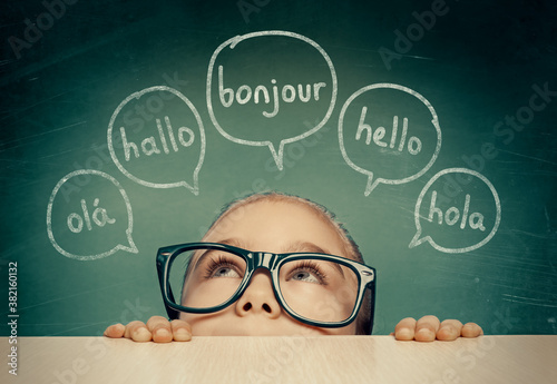 Beautiful cute little girl with eyeglasses hiding under table and looking at hello word in french, english, spanish, portuguese and german in speech balloons. K-12 foreign language learning concept.