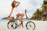 young attractive smiling woman in white dress riding on tropical beach on bicycle sunglasses traveling on summer vacation in Thailand, boho fashion style