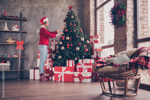 Photo of lovely young girl hands hang toys evergreen tree wear santa headwear red sweater decorated living room indoors