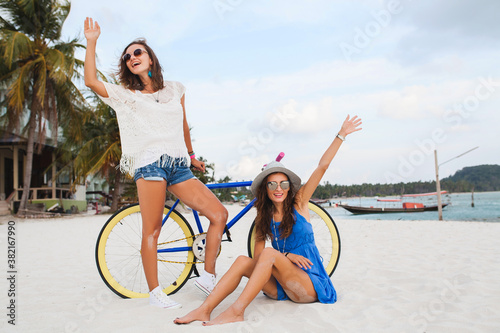 happy female friends having fun on tropical beach  women traveling on vacation in Thailand with bicycle  summer fashion trend